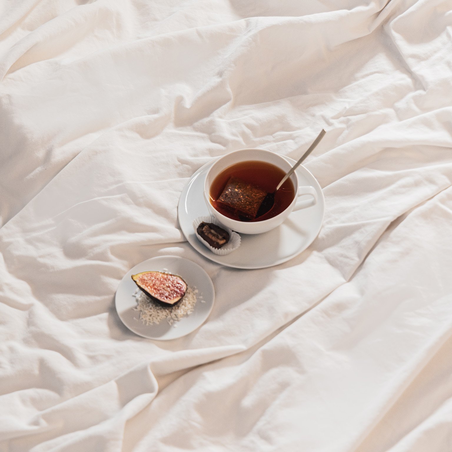 TAC tea cup filled with tea, saucer and bread plate with a fig stand on a bed sheet.